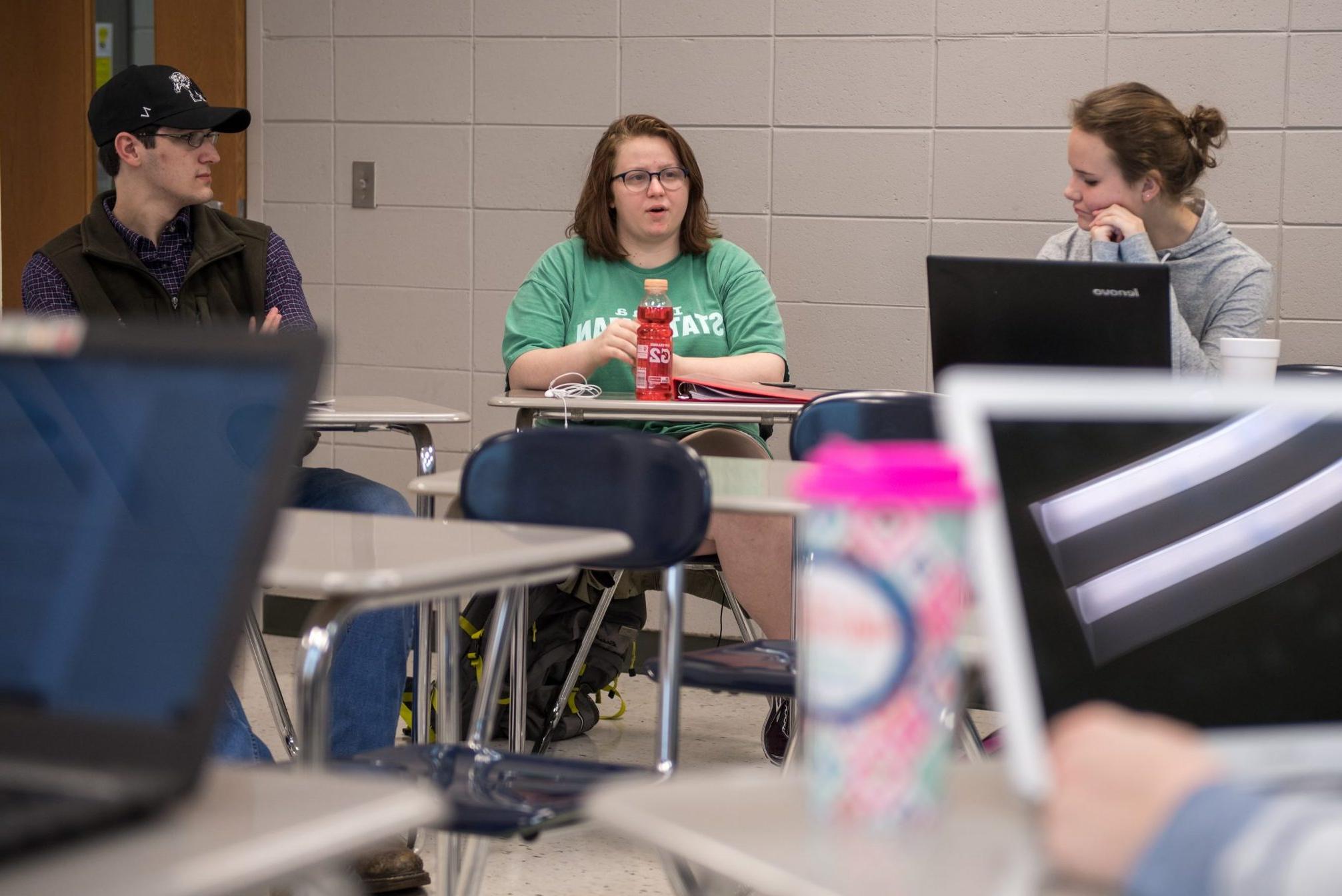 一个学生坐在课桌上说话，另外两个学生在听, 她的两边各一个.